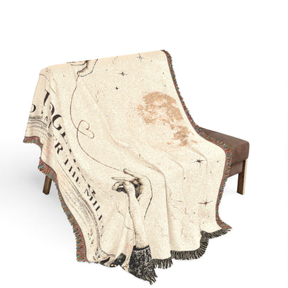 A cream-colored woven throw blanket with fringe, draped over a brown armchair. The blanket features a vintage-style celestial map design with black script text, star illustrations, and a brown celestial body graphic.