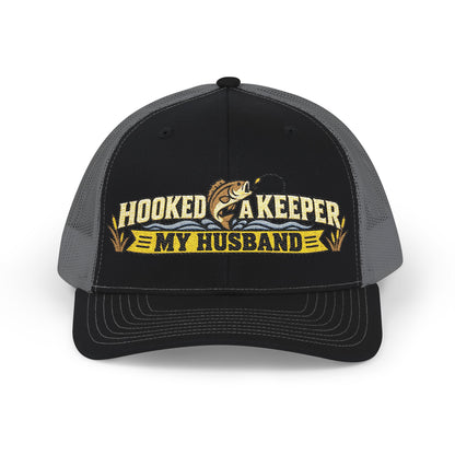 A black and gray trucker hat with a front graphic that reads 'HOOKED A KEEPER MY HUSBAND' in yellow and white, featuring a fishing rod and fish illustration.