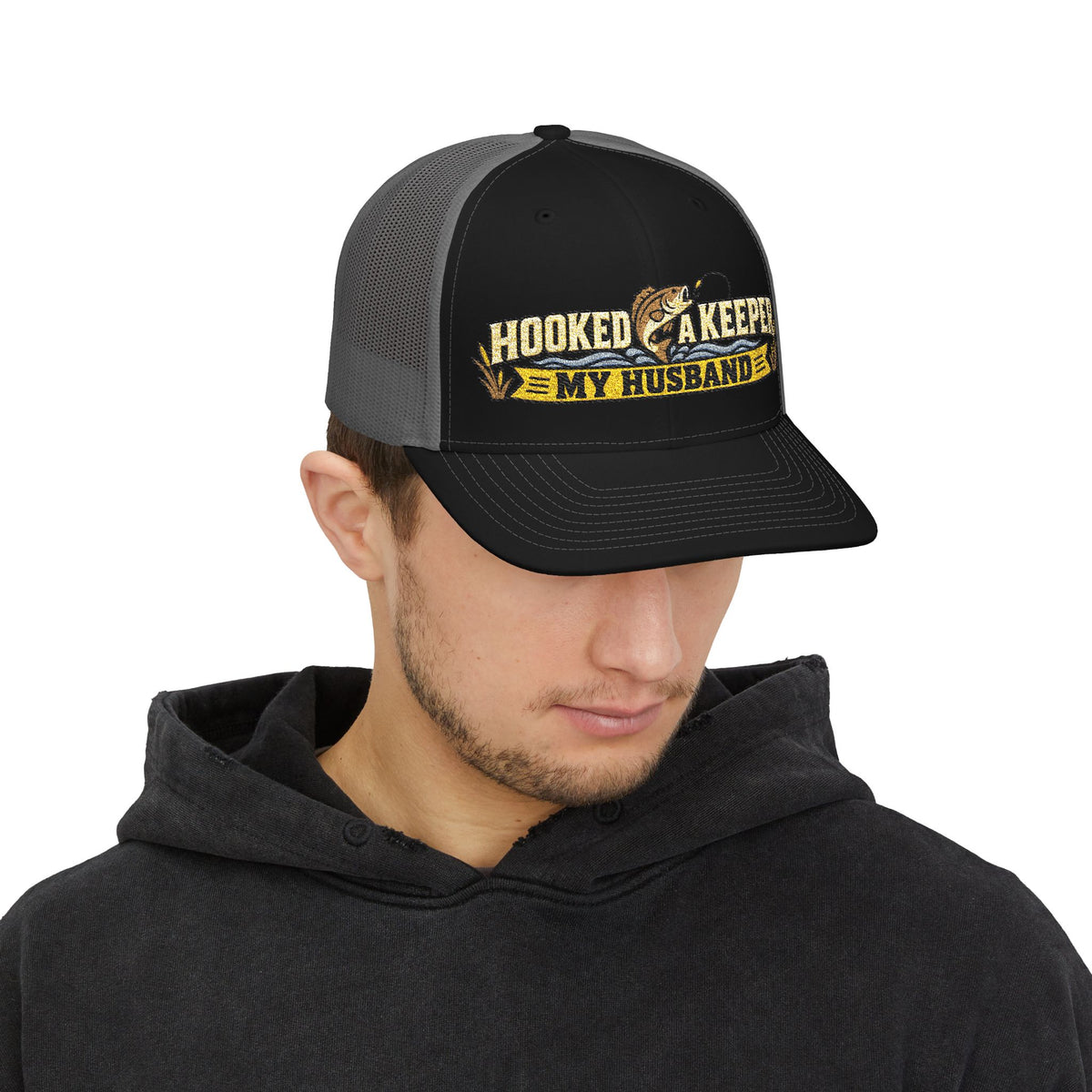 A black and gray trucker hat with a front graphic that reads 'HOOKED A KEEPER MY HUSBAND' in yellow and white, featuring a fishing rod and fish illustration.