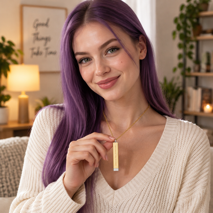 A woman with vibrant purple hair wearing a cream knit sweater and a gold pendant necklace, holding the pendant in a cozy, warmly lit living room setting.