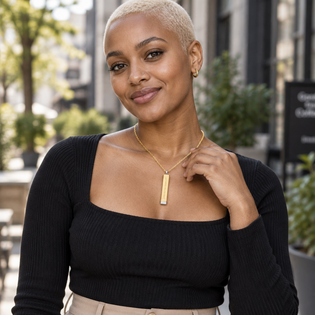 A woman with a short blonde haircut wearing a black ribbed long-sleeve top and a gold bar pendant necklace, posing outdoors with a blurred urban background.