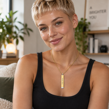 A woman with short blonde hair wearing a black ribbed tank top and a gold bar necklace with a vertical engraved plate, posing in a brightly lit, modern living room setting.