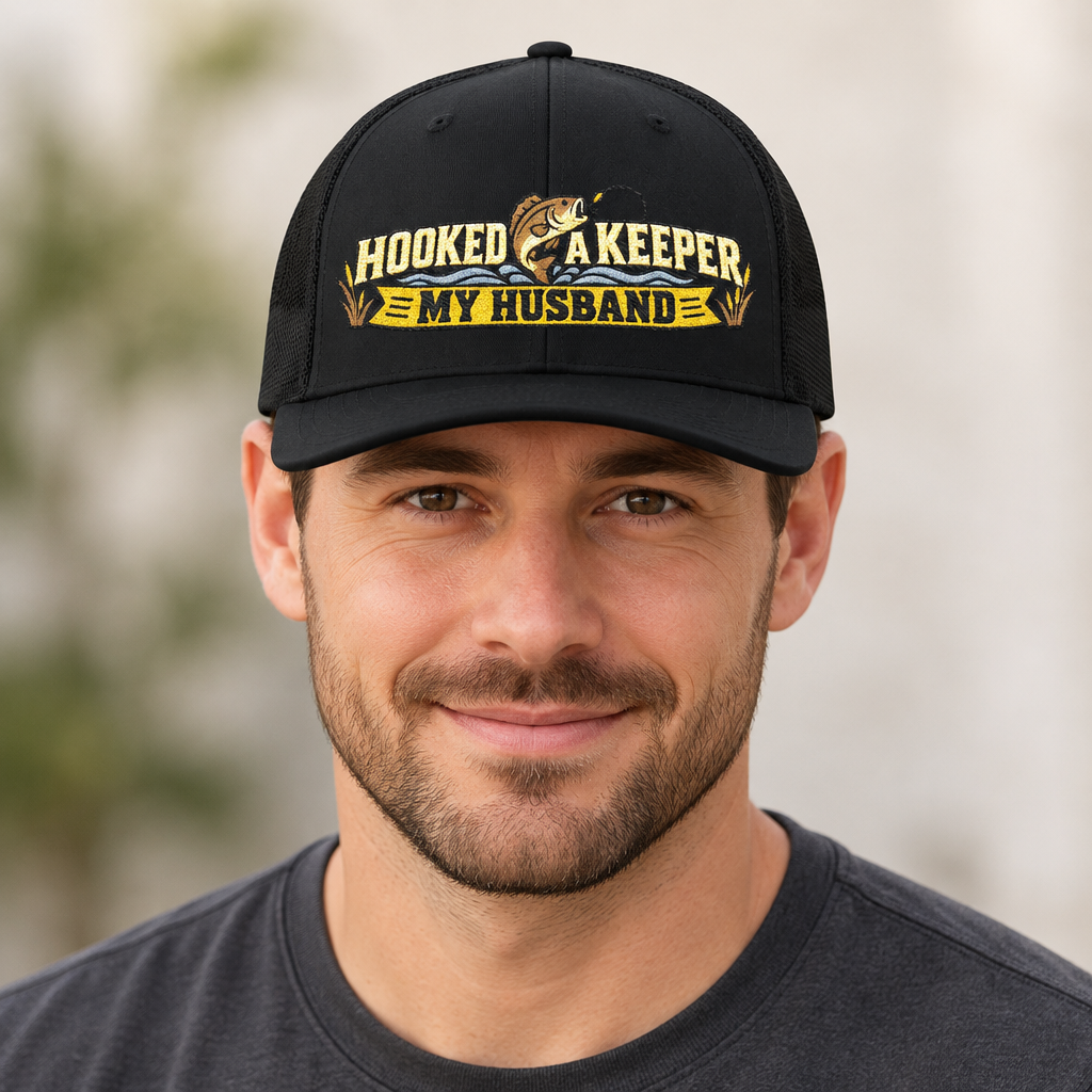 A man wearing a black baseball cap with a yellow and gold graphic that reads 'HOOKED A KEEPER MY HUSBAND' with a fish illustration, against a softly blurred outdoor background.