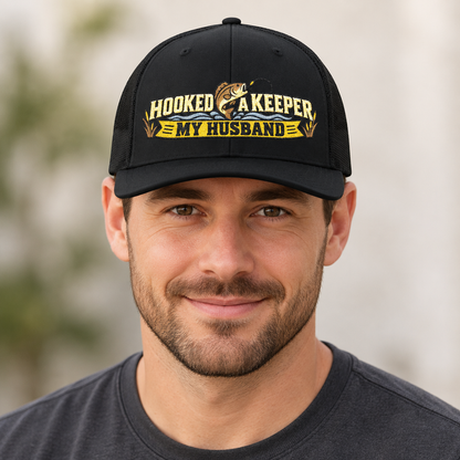 A man wearing a black baseball cap with a yellow and gold graphic that reads 'HOOKED A KEEPER MY HUSBAND' with a fish illustration, against a softly blurred outdoor background.