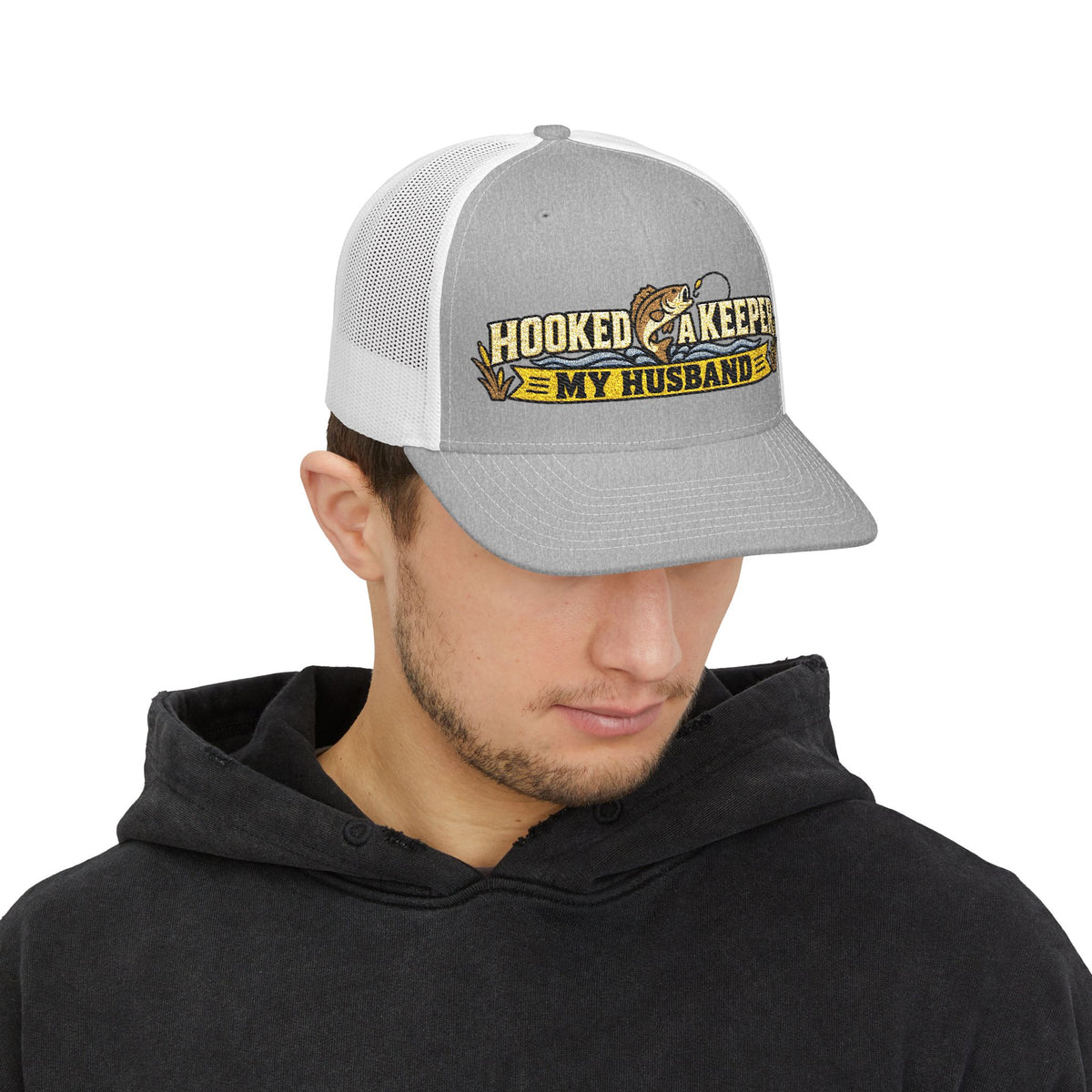 A man wearing a gray and white trucker hat with a graphic that reads 'HOOKED LIKE A KEEPER MY HUSBAND' in yellow, black, and brown, paired with a black hoodie against a white background.