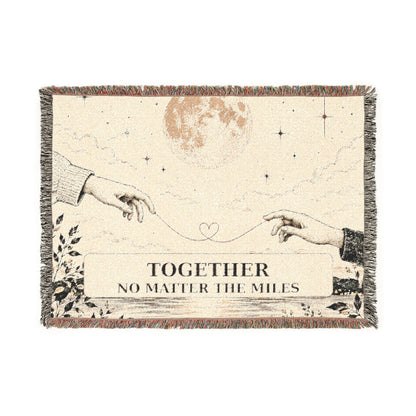 A rectangular woven throw blanket with fringe edges, featuring an illustrated celestial scene with two reaching hands, stars, a moon, and the text 'TOGETHER NO MATTER THE MILES' on a cream background.