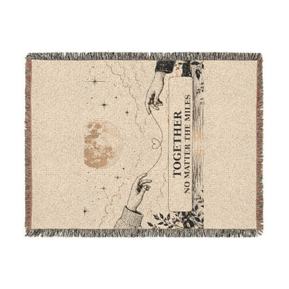 A rectangular woven throw blanket with fringe on all sides, featuring a beige background and a printed design of two reaching hands, stars, a moon, and the text 'TOGETHER NO MATTER THE MILES'.