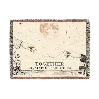 A square woven throw blanket with fringe edges, featuring an illustration of two hands reaching toward a heart, a moon, stars, and the text 'TOGETHER NO MATTER THE MILES' on a cream background.