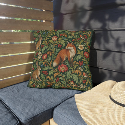 A square decorative throw pillow with a detailed woodland pattern of a fox, rabbits, and flowers on a dark green background, styled on a blue outdoor patio cushion next to a straw hat.
