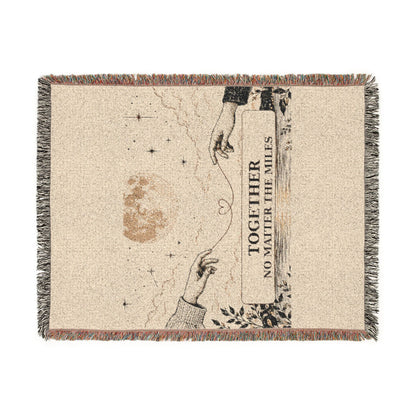 A rectangular woven throw blanket with fringed edges, featuring a beige textured background and a printed illustration of two reaching hands, stars, a moon, and the text 'TOGETHER NO MATTER THE MILES'.
