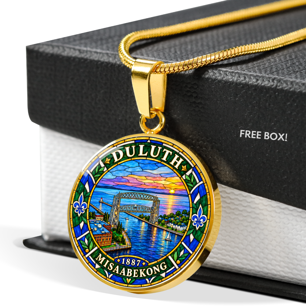 A gold-tone circular pendant necklace featuring a colorful stained-glass-style Duluth, Minnesota design with the words 'DULUTH' and 'MISAAABEKONG' and '1887', displayed on a black gift box with the text 'FREE BOX!'.