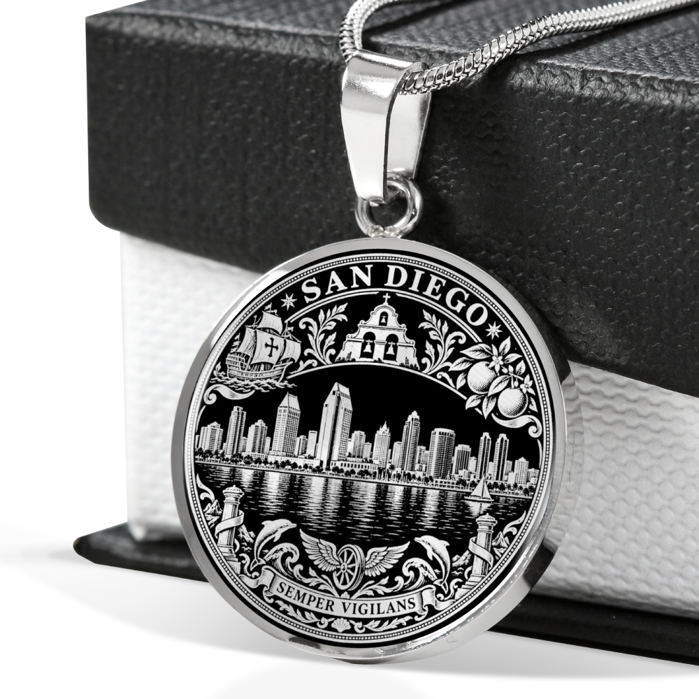 A round silver-toned pendant necklace featuring an engraved San Diego cityscape and decorative border, hanging from a chain in front of a black gift box.