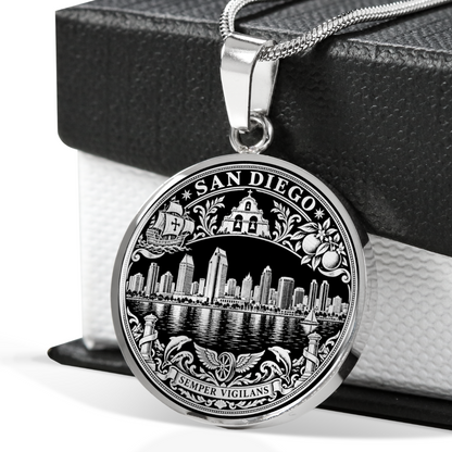 A round silver-toned pendant necklace featuring an engraved San Diego cityscape and decorative border, hanging from a chain in front of a black gift box.