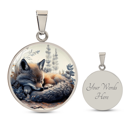 Sleeping fox necklace featuring a curled-up red fox with warm orange fur among soft blue-grey woodland foliage on a circular pendant with engraving option