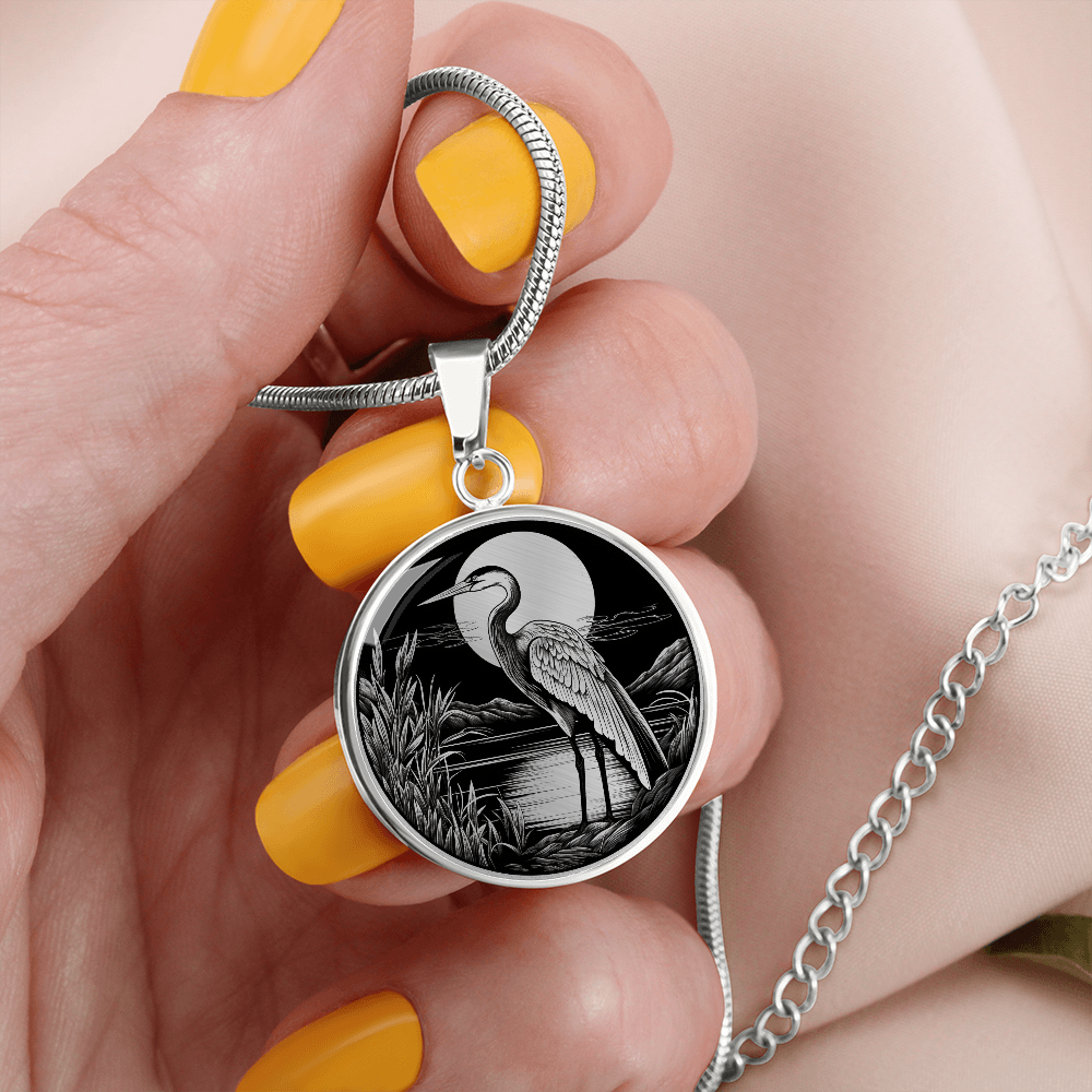 Heron necklace featuring a long-legged wading bird beside a marsh with reeds and trees in detailed black-and-white artwork on a round pendant.