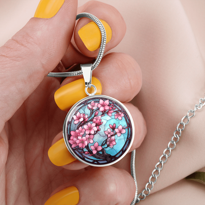 Woman holding a Cherry blossom necklace featuring pink sakura flowers on dark branches against a turquoise-blue stained-glass-style circular pendant.