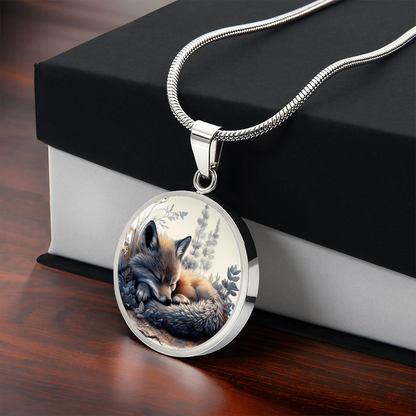 Sleeping fox necklace featuring a curled-up red fox with warm orange fur among soft blue-grey woodland foliage on a circular pendant with gift box
