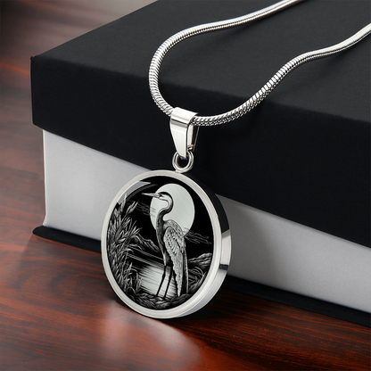 Heron necklace featuring a long-legged wading bird beside a marsh with reeds and trees in detailed black-and-white artwork on a round pendant.