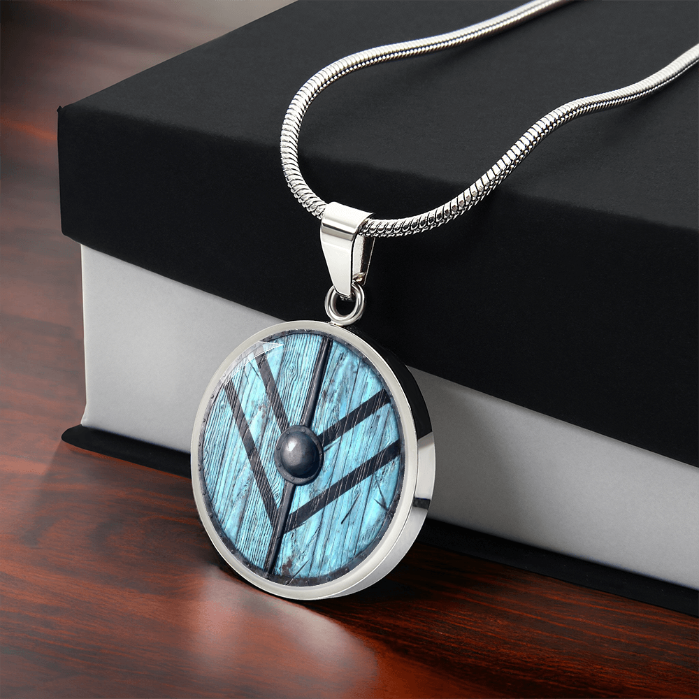 Viking shield necklace featuring a weathered turquoise-blue round shield with dark reinforcing bands and central metal boss on a circular pendant with gift box