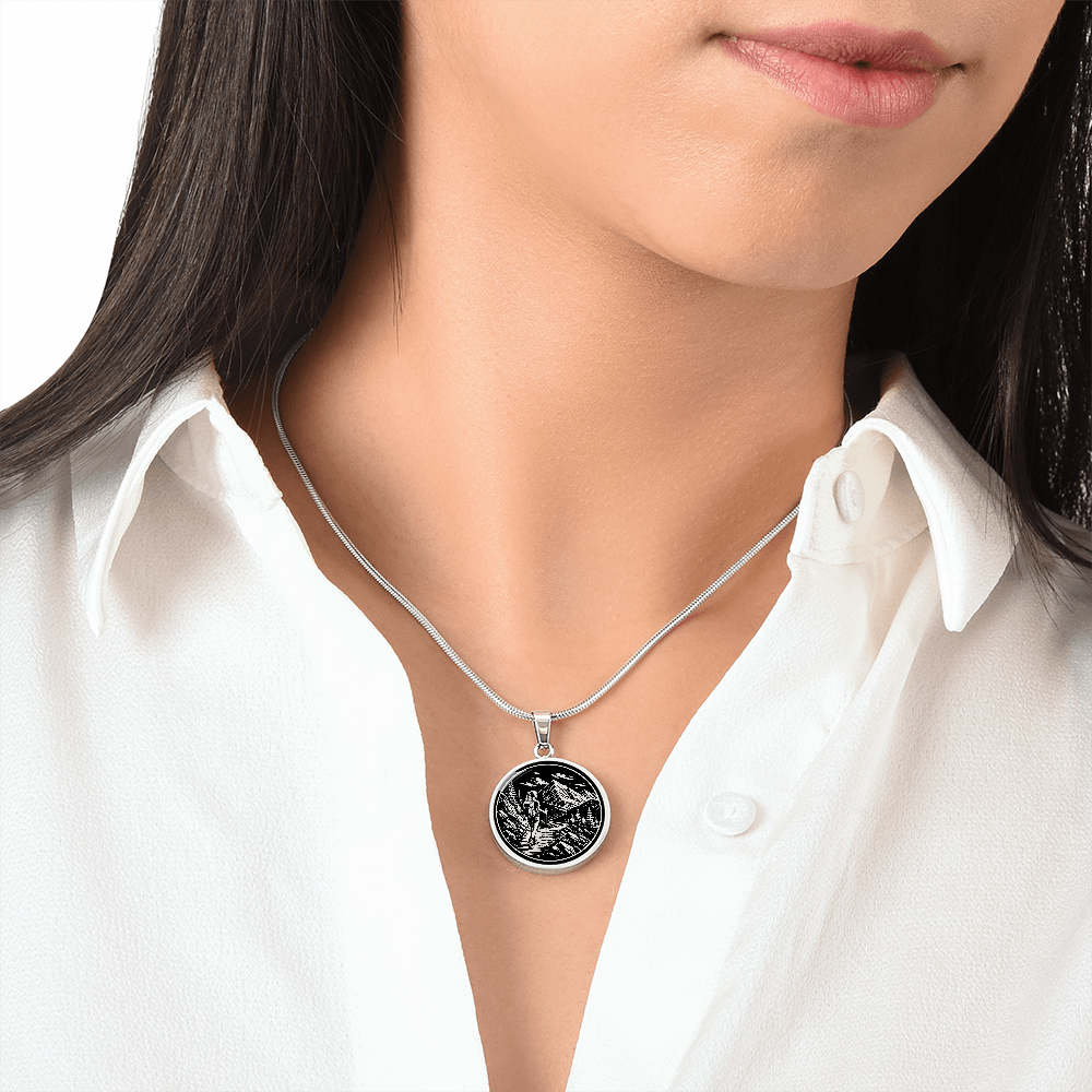 Silver-colored hiking pendant with a lone woman carrying a backpack and trekking pole beneath mountain peaks and evergreen trees.