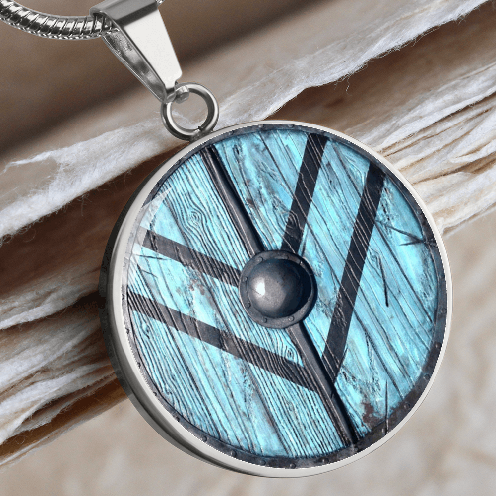 Viking shield necklace featuring a weathered turquoise-blue round shield with dark reinforcing bands and central metal boss on a circular pendant.