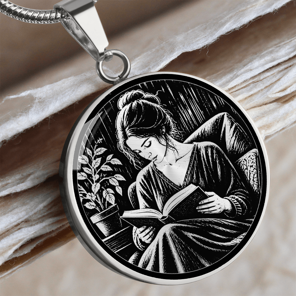 Woman reading necklace featuring linocut-style artwork of a woman reading an open book in a cushioned chair beside a leafy plant.
