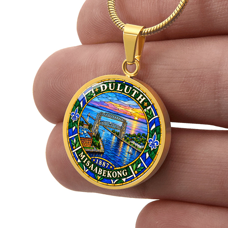 Male hand holding a Gold finish Duluth harbor pendant necklace showing the Aerial Lift Bridge, blue lake water, sunset, fleur-de-lis motifs and Duluth lettering