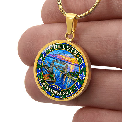 Male hand holding a Gold finish Duluth harbor pendant necklace showing the Aerial Lift Bridge, blue lake water, sunset, fleur-de-lis motifs and Duluth lettering