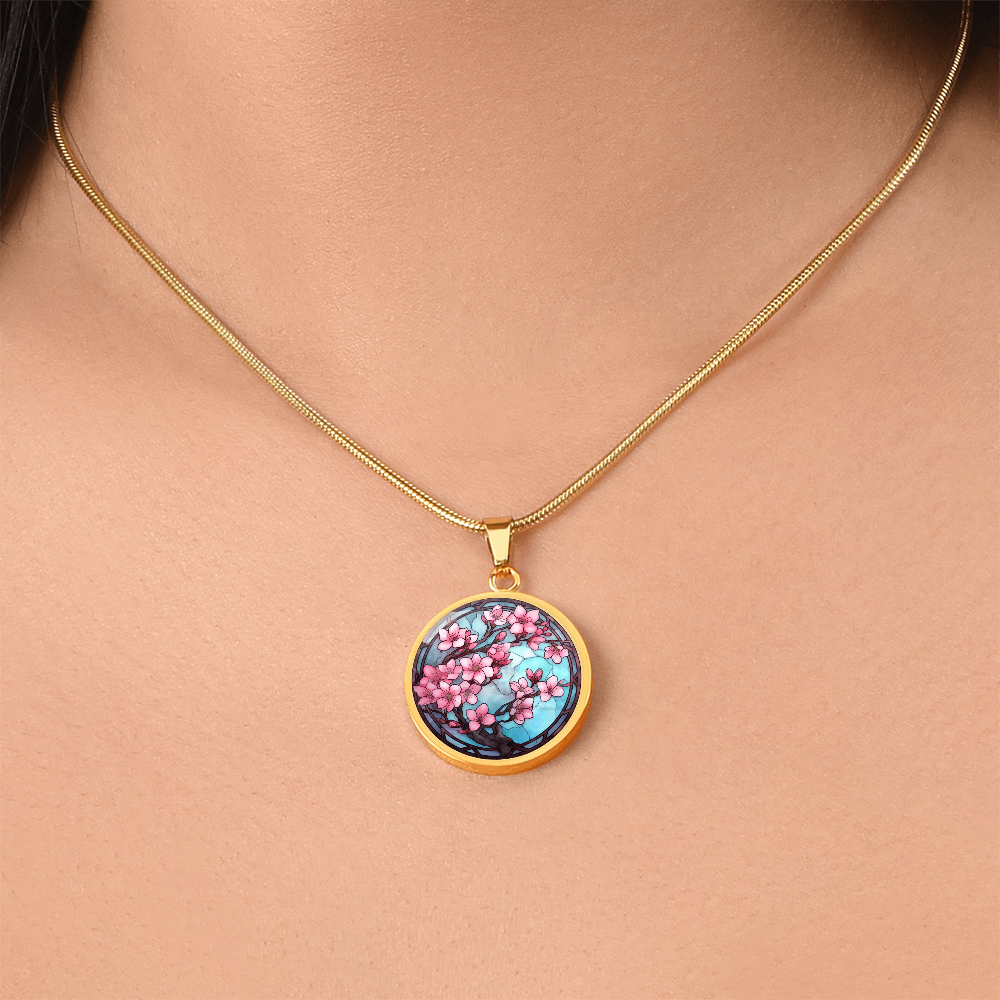 Woman wearinga gold Cherry blossom necklace featuring pink sakura flowers on dark branches against a turquoise-blue stained-glass-style circular pendant.