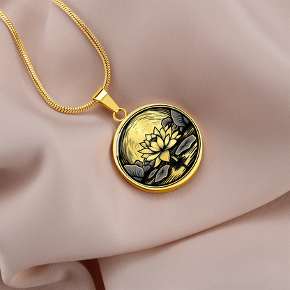 Gold finish lotus flower necklace featuring vintage linocut-style artwork of a blossom rising above a reflective pond on a cream colored linen.