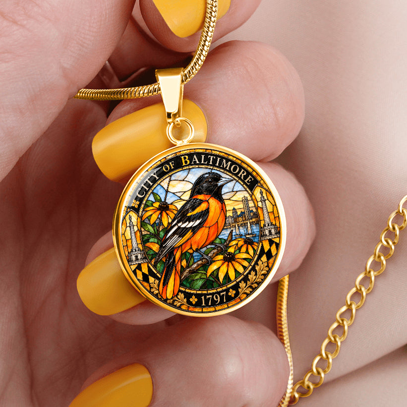 Woman holding a gold Baltimore Oriole stained glass pendant necklace on silver chain with colorful bird, floral design and Baltimore city heritage border