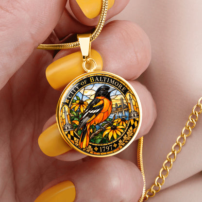Woman holding a gold Baltimore Oriole stained glass pendant necklace on silver chain with colorful bird, floral design and Baltimore city heritage border