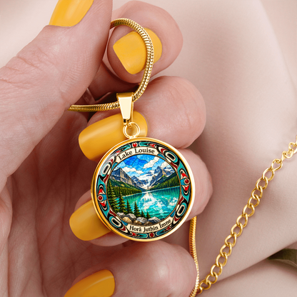 Woman holding a Gold finish Lake Louise pendant necklace showing bright blue mountain lake, snowy peaks, pine trees and decorative stained-glass artwork