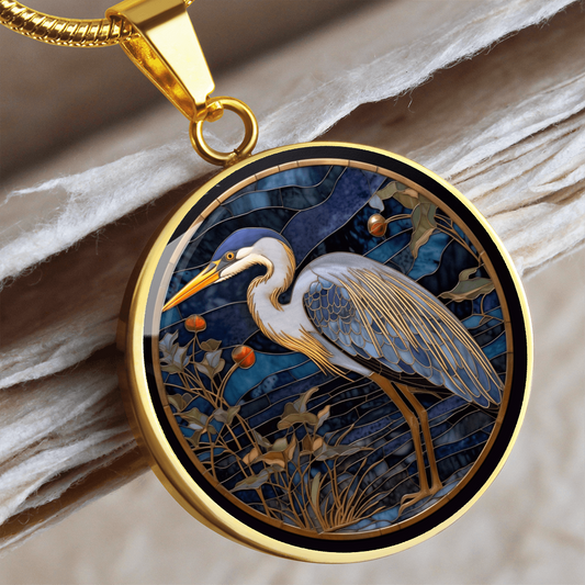 Cloisonné-style heron necklace featuring a blue-grey heron with orange beak, gold-outlined feathers and foliage against a deep navy background.