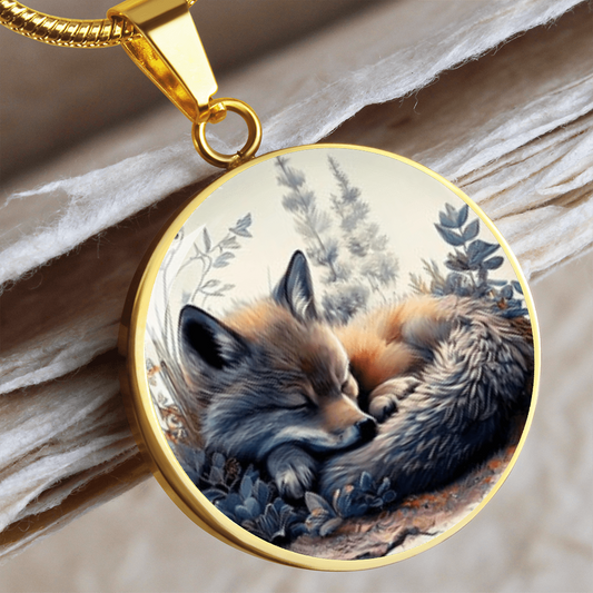 Sleeping fox necklace featuring a curled-up red fox with warm orange fur among soft blue-grey woodland foliage on a circular pendant.