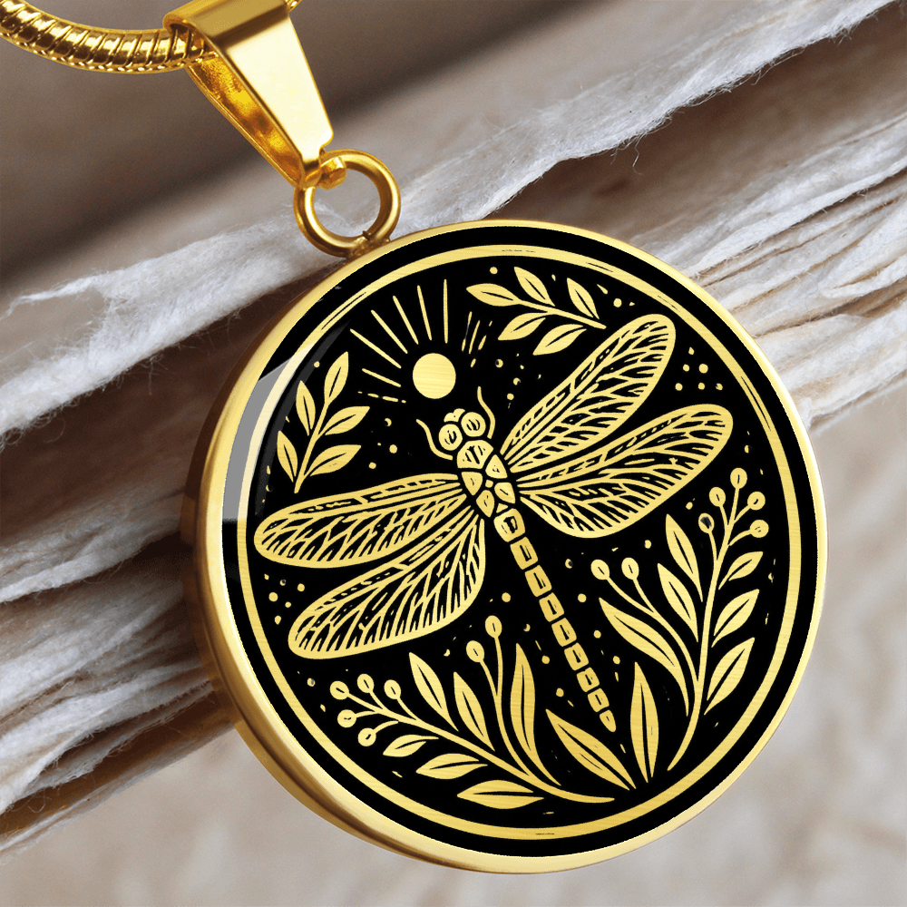Dragonfly necklace with linocut-style artwork showing four patterned wings, botanical leaves and sun rays on a black circular pendant.