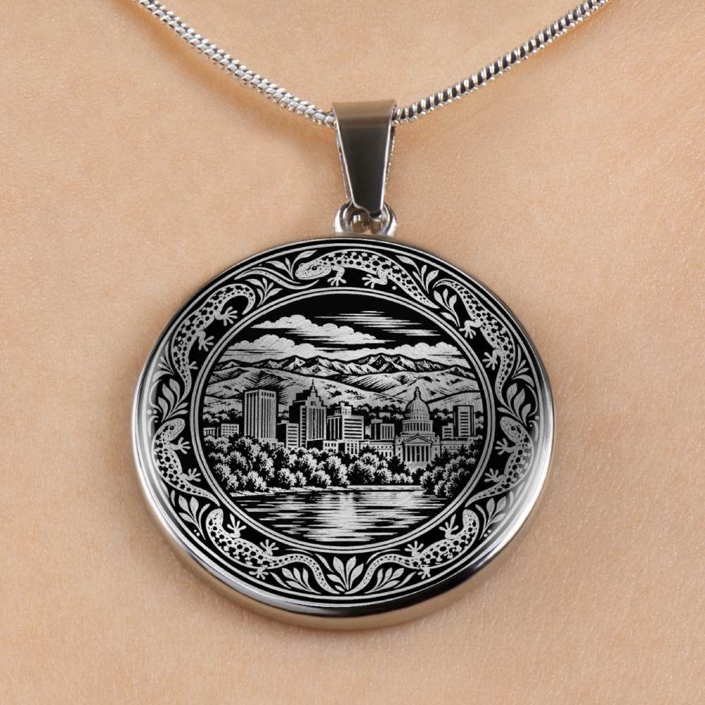 A silver oval pendant necklace featuring an engraved black and white cityscape of boise, Idaho and mountain scene, shown worn on a person's neck against a light beige skin background.
