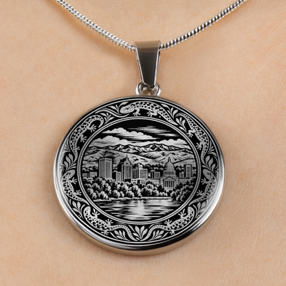A silver oval pendant necklace featuring an engraved black and white cityscape of boise, Idaho and mountain scene, shown worn on a person's neck against a light beige skin background.