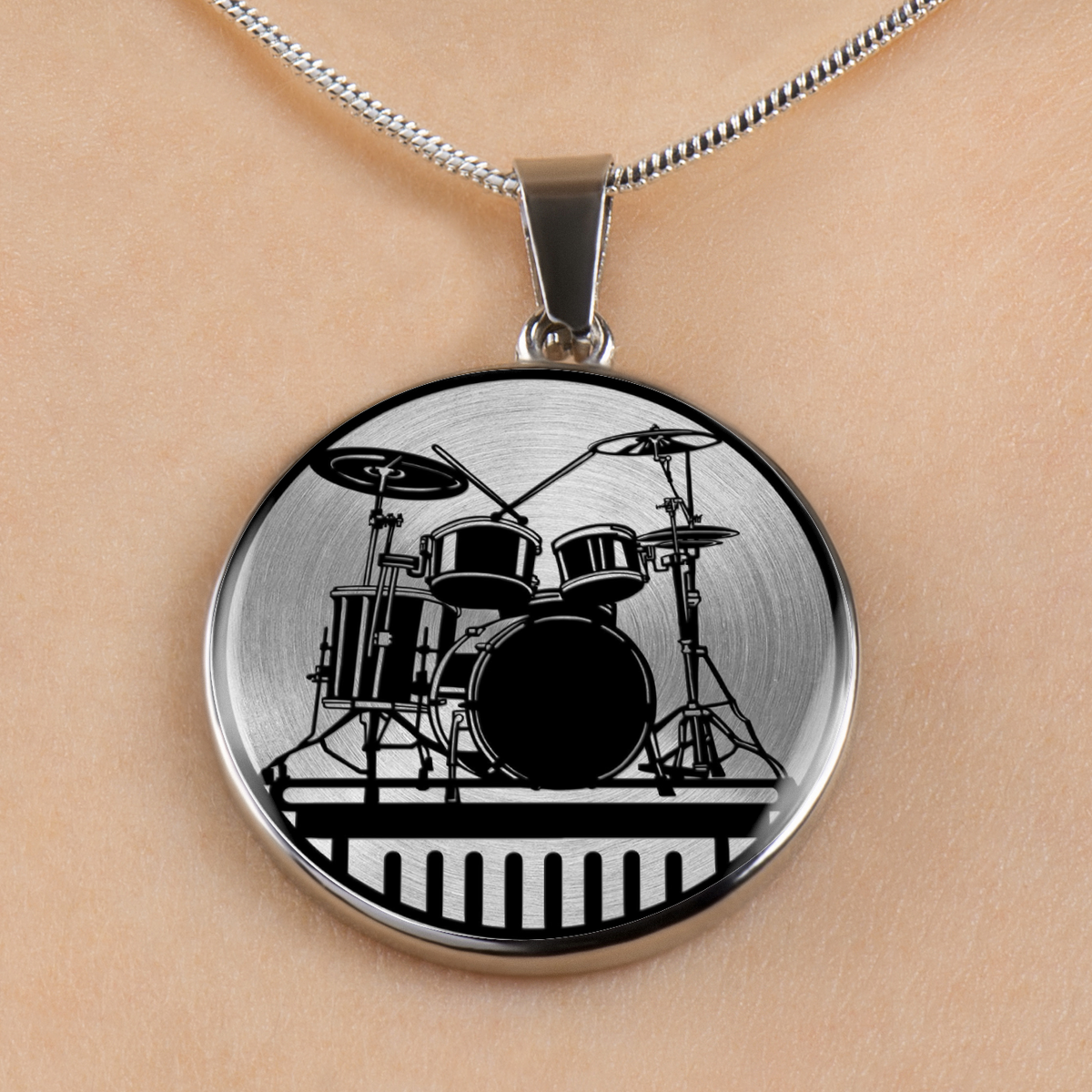 A silver round pendant necklace featuring a black graphic of a drum set, shown worn on a person's neck against a light skin tone background.