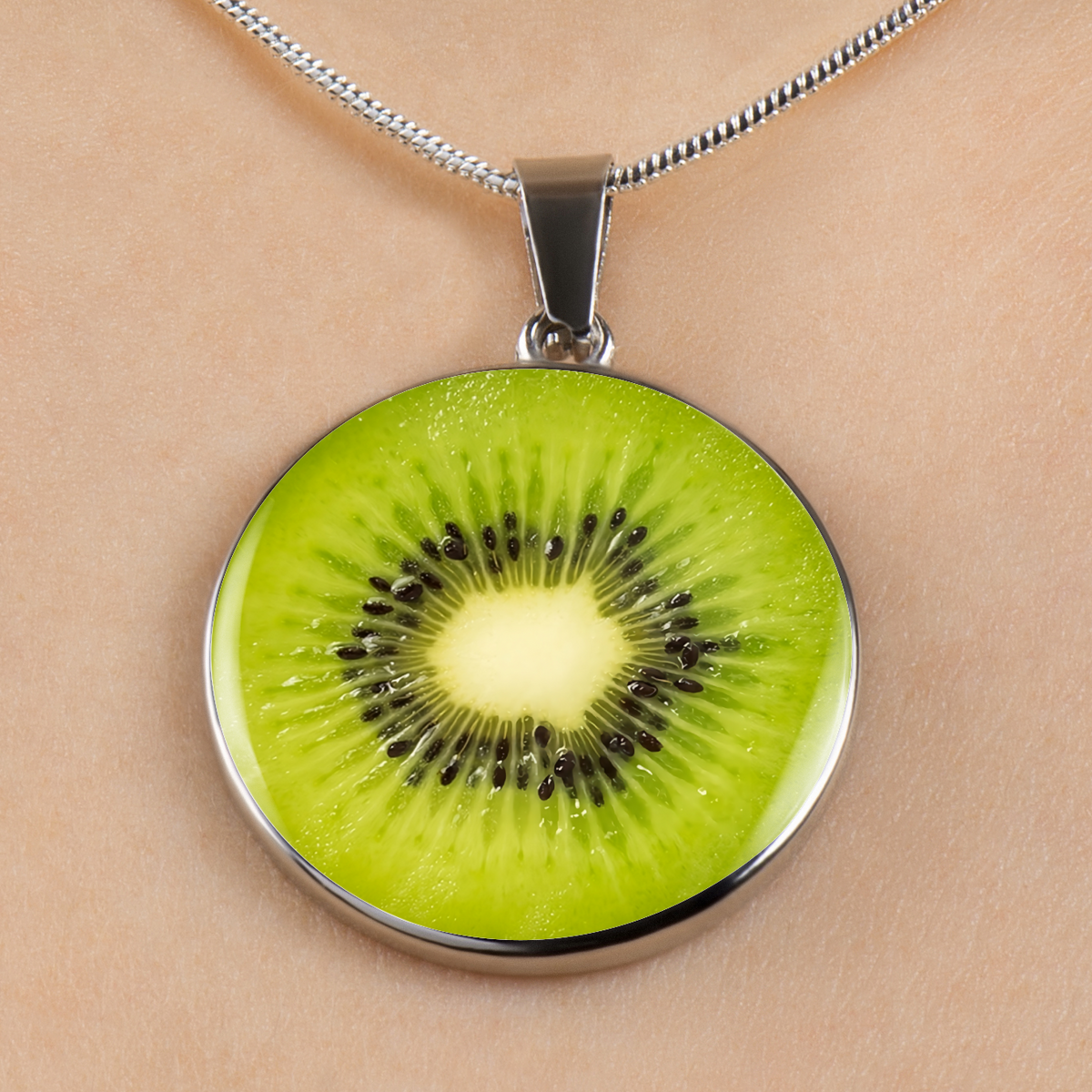 A round pendant necklace featuring a realistic kiwi slice design with bright green flesh, black seeds, and a pale yellow center, set in a silver-toned metal frame and hanging from a silver chain against a light beige skin background.