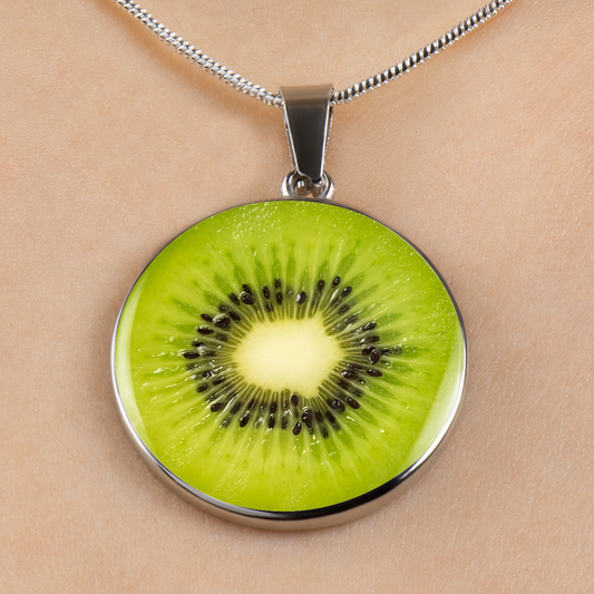 A round pendant necklace featuring a realistic kiwi slice design with bright green flesh, black seeds, and a pale yellow center, set in a silver-toned metal frame and hanging from a silver chain against a light beige skin background.