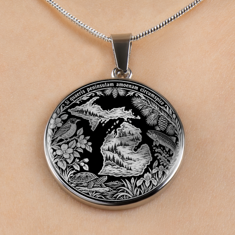 A silver oval pendant necklace featuring an engraved design of the Great Lakes Michigan shoreline, complete with a fish, floral details, and the Latin inscription 'S. maris peninsulae amoenam circumspice'.