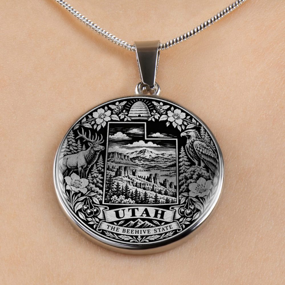 A silver oval pendant necklace featuring an engraved Utah state seal with a mountain landscape, deer, beehive, flowers, and the words 'UTAH THE BEEHIVE STATE,' shown on a chain against a light skin-toned background.